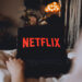 Netflix logo on the laptop screen. Young woman watching netflix channel sitting at home in the evening.